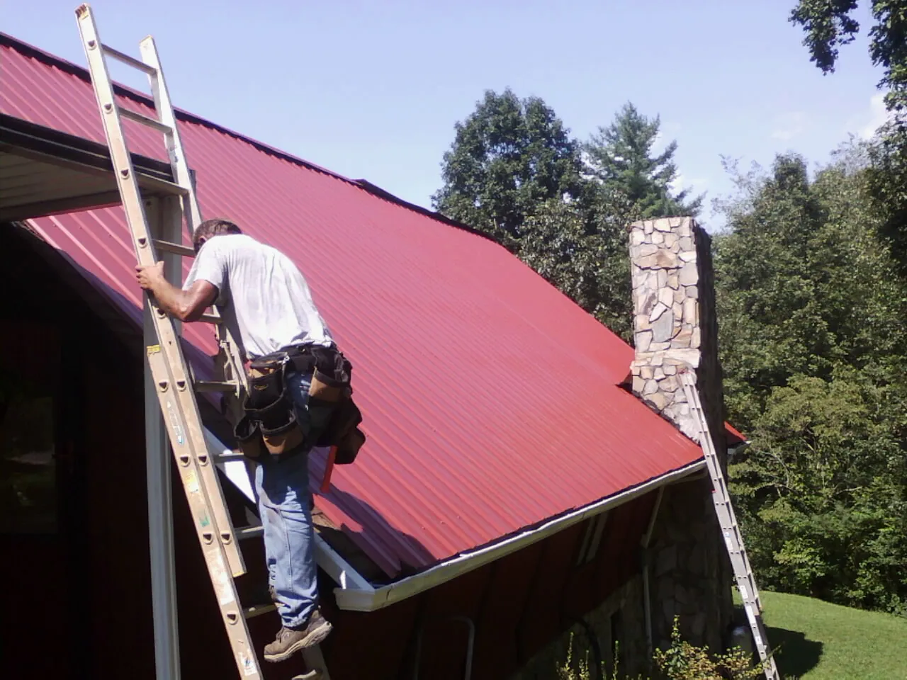 Roofer on a ladder beside a red metal roof and stone chimney — Soffit & Fascia Repair by Roofing Brothers in Connellsville